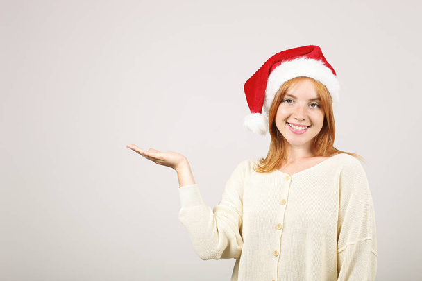 Close up portrait of beautiful redheaded young woman wearing Santa Claus hat & white sweater with satisfied facial expression. Cute female celebrating New Year. Copy space, isolated on gray background - Photo, Image