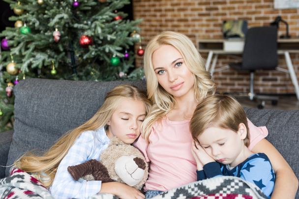 νεαρή μητέρα βλέπει κάμερα ενώ κάθεται με χαριτωμένο μικρό chikdren στο πιτζάμες στον ύπνο στον καναπέ στο χρόνο Χριστουγέννων - Φωτογραφία, εικόνα
