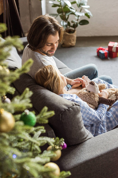 high angle view of happy father looking at cute daughter in pajamas holding teddy bear and sleeping on couch at christmas time - Valokuva, kuva