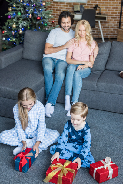 high angle view of adorable children in pajamas opening christmas gifts while happy parents sitting behind  - Foto, Bild
