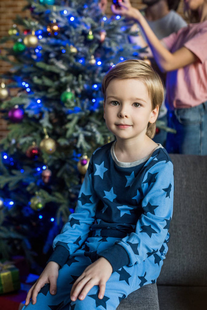 adorable niño en pijama sentado en el sofá y sonriendo a la cámara en Navidad
 - Foto, Imagen