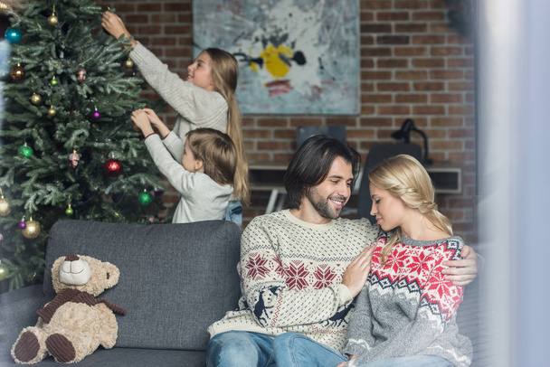 selective focus of happy parents sitting in sofa while children decorating christmas tree behind  - Fotoğraf, Görsel