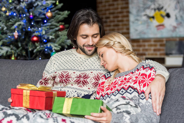 beautiful young couple holding christmas gifts and sitting together on couch - Photo, image