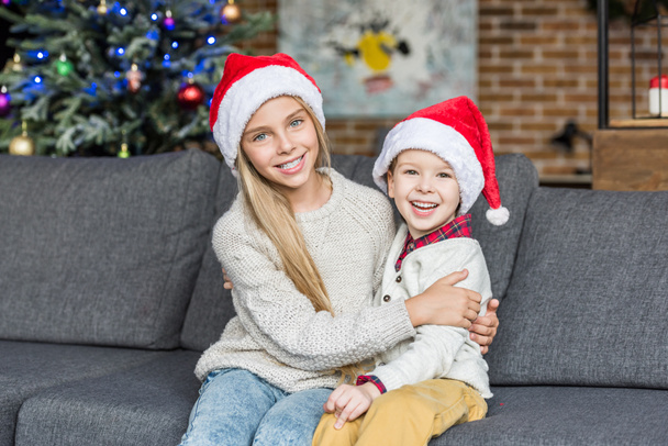 süße, glückliche Kinder in Weihnachtsmützen, die zusammen auf der Couch sitzen und in die Kamera lächeln  - Foto, Bild