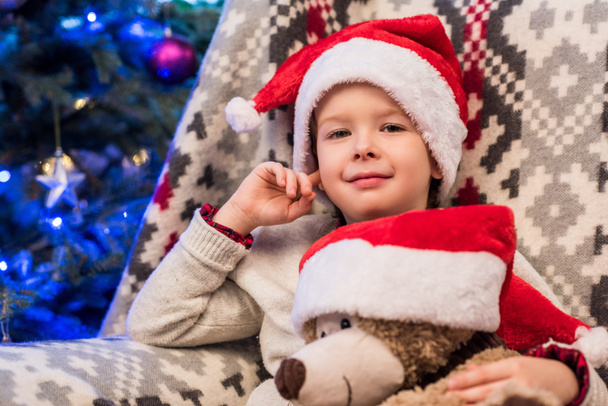 lindo niño en santa sombrero sosteniendo osito de peluche y sonriendo a la cámara en Navidad
  - Foto, imagen