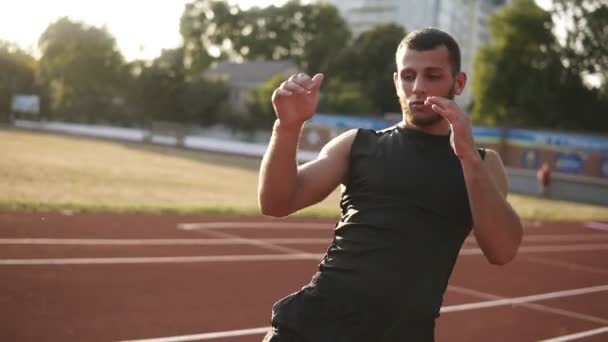 Muscular athlete young male stretching his legs on a running track in stadium, preparing for training. Lift his legs up. Sun shines - Filmagem, Vídeo