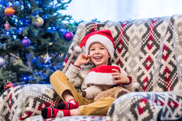 happy little boy holding teddy bear in santa hat and smiling at camera   - Фото, зображення