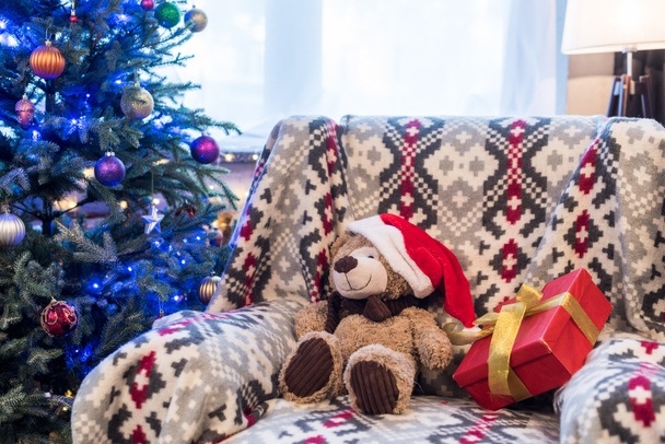 oso de peluche en sombrero de santa y caja de regalo en sofá y árbol de Navidad decorado detrás
 - Foto, Imagen