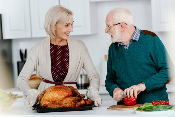 happy senior couple cooking thanksgiving turkey together and looking at each other - Foto, Imagen
