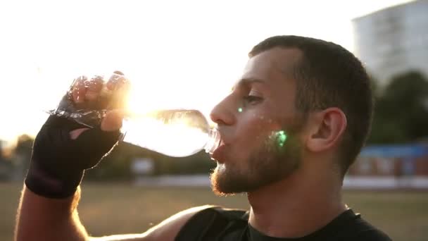 Side view of a young male boxer in black bandages has a rest during the training outdoors, drinking water from a plastic bottle. Sun shines on the background. - Filmati, video