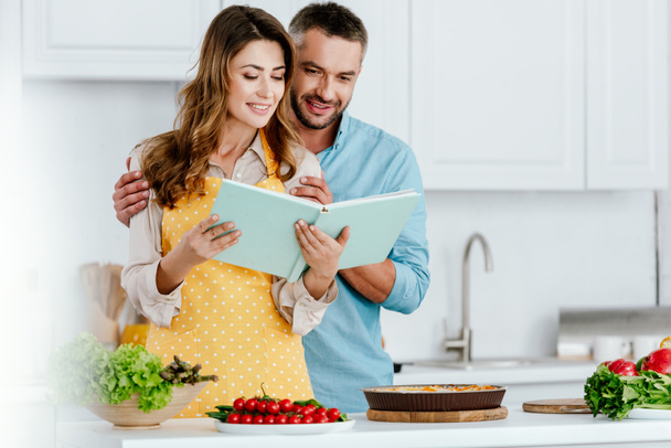 happy couple reading recipe book while cooking together at kitchen - Fotó, kép