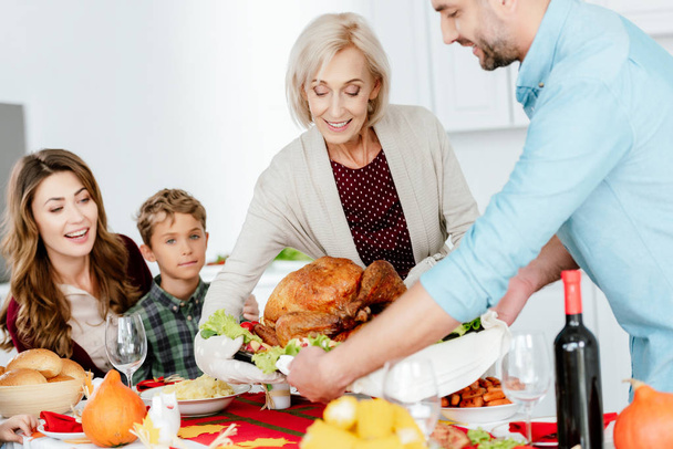 счастливая пожилая женщина и взрослый мужчина, несущий запеченную индейку на ужин в честь Дня Благодарения с семьей
  - Фото, изображение