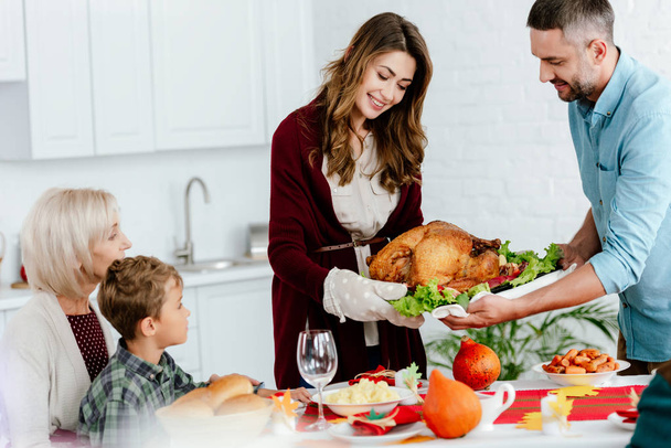 улыбающаяся взрослая пара, несущая запечённую индейку на ужин в честь Дня Благодарения с большой семьей
  - Фото, изображение