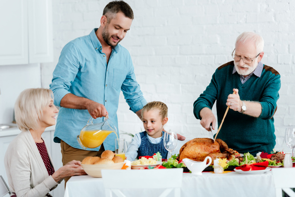 abuelo rebanando pavo mientras el hombre adulto vierte jugo en la mesa servida para la celebración de Acción de Gracias con la familia
  - Foto, Imagen