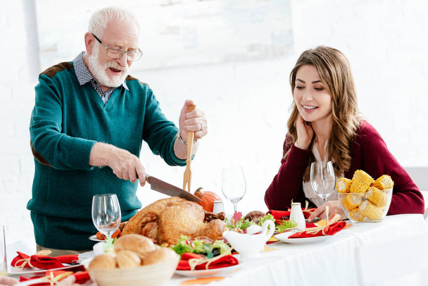 happy senior man slicing delicious turkey for thanksgiving celebration while his adult daughter sitting near at table  - Photo, Image