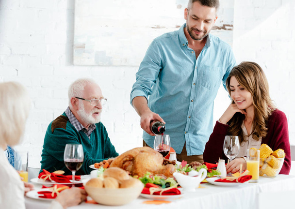 Erwachsener Mann gießt Wein ins Glas am Serviertisch, während Familie zu Hause Erntedank feiert - Foto, Bild