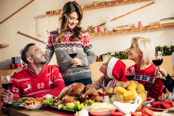 όμορφη χαμογελαστή γυναίκα με ποτήρι κρασιού κάνοντας πρόποση κατά τη διάρκεια των Χριστουγέννων δείπνο με ευτυχισμένη οικογένεια στο σπίτι  - Φωτογραφία, εικόνα