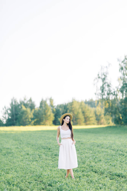 Girl walking on the field, in a hat and summer dress. Smiling and laughing, beautiful sunset in the forest and in nature. White dress and rye, sloping fields. Happy traveler, lifestyle. - Photo, image