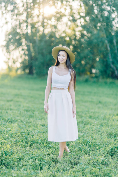 Girl walking on the field, in a hat and summer dress. Smiling and laughing, beautiful sunset in the forest and in nature. White dress and rye, sloping fields. Happy traveler, lifestyle. - Fotó, kép