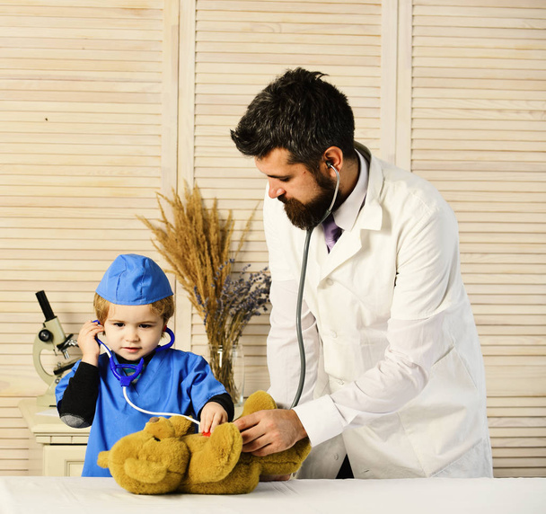 Vet and little assistant examine teddy bear. Father and kid with attentive faces playing doctor. Healthcare and childhood concept. Man and boy hold stethoscope on wooden background. - Photo, Image