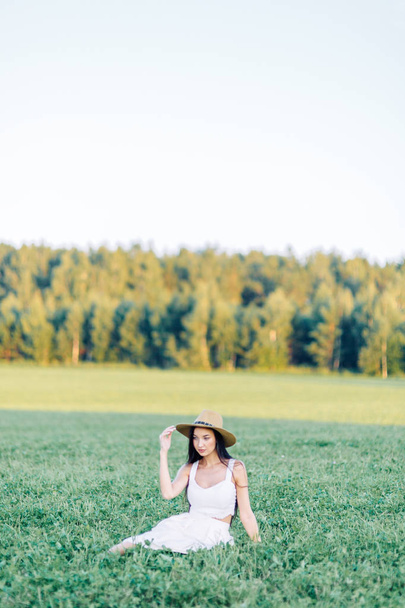 Chica caminando en el campo, con un sombrero y vestido de verano. Sonriendo y riendo, hermoso atardecer en el bosque y en la naturaleza. Vestido blanco y centeno, campos inclinados. Viajero feliz, estilo de vida
. - Foto, imagen