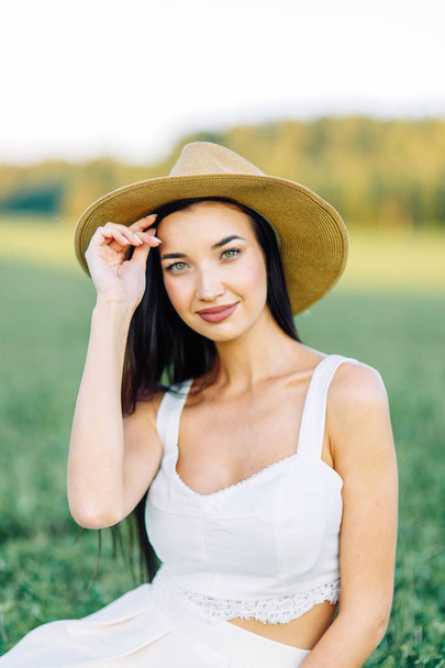 Girl walking on the field, in a hat and summer dress. Smiling and laughing, beautiful sunset in the forest and in nature. White dress and rye, sloping fields. Happy traveler, lifestyle. - Foto, Bild