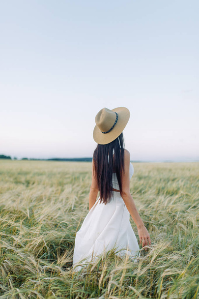 Κοπέλα περπάτημα στο πεδίο, σε ένα καπέλο και το καλοκαίρι φόρεμα. Χαμογελαστός και γελά, όμορφο ηλιοβασίλεμα στο δάσος και στη φύση. Λευκό φόρεμα και σίκαλη, επικλινές πεδία. Ευτυχισμένος ταξιδιώτης, lifestyle. - Φωτογραφία, εικόνα