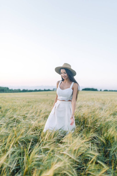 Lány séta a mező, a kalap és a nyári ruha. Mosolyogva és nevetve, gyönyörű naplemente az erdőben, és a természetben. Fehér ruha és a rozs, a lejtős területeken. Boldog utazó, életmód. - Fotó, kép