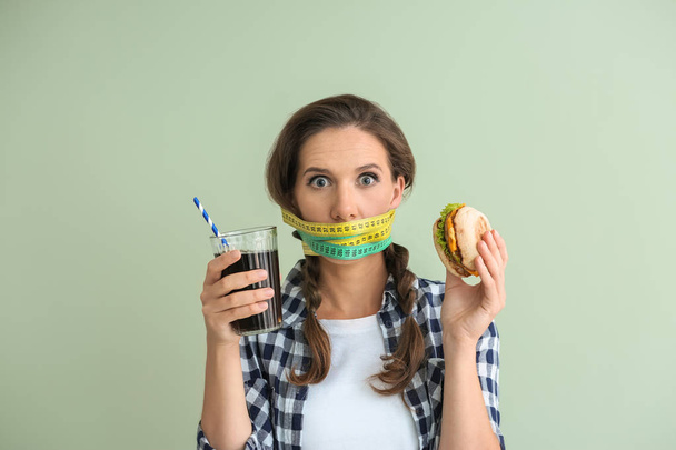 Emotional woman with measuring tapes around her mouth and unhealthy food on color background. Diet concept - Photo, image