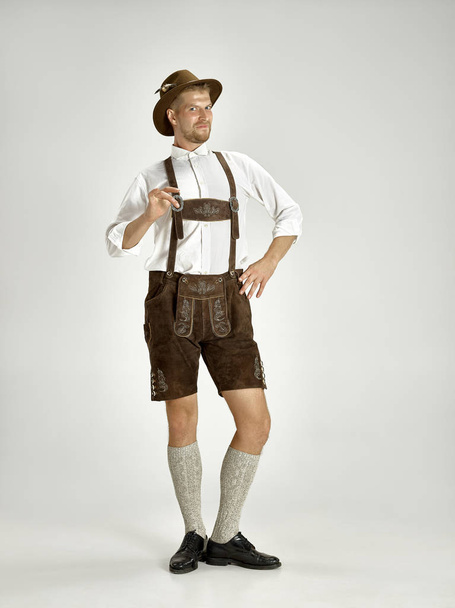 Portrait of Oktoberfest young man in hat, wearing a traditional Bavarian clothes standing at full-length at studio. The celebration, oktoberfest, festival concept - Photo, image