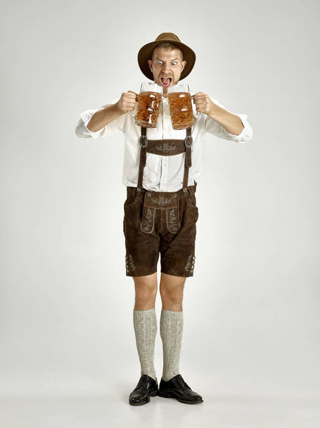 Portrait of Oktoberfest young man in hat, wearing a traditional Bavarian clothes standing with beer at full-length at studio. The celebration, oktoberfest, festival concept - Фото, зображення