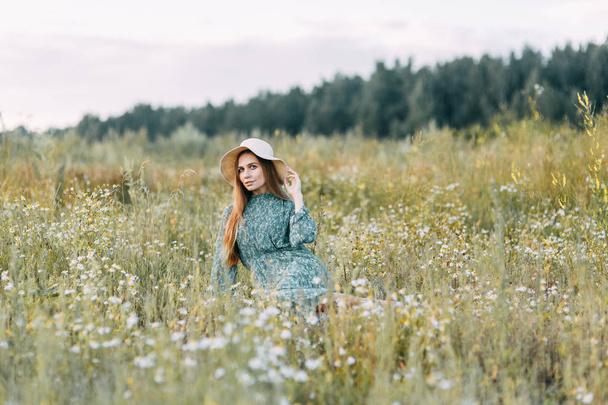 Beautiful Russian girl in a hat and summer dress, walking on the field in the grass to the waist. Happy traveler, collecting herbs, smiling. Sunset in the forest. - Foto, imagen