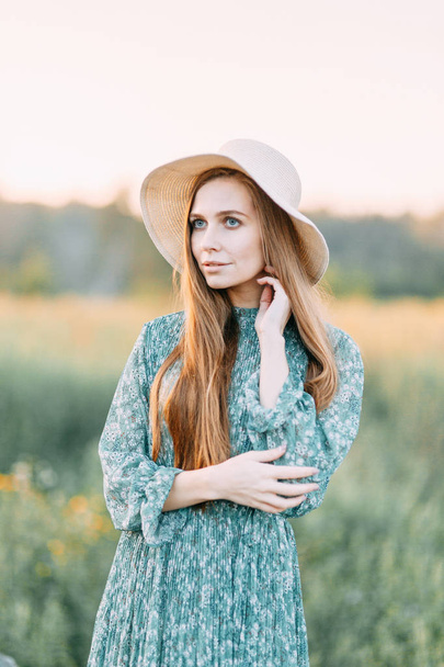 Beautiful Russian girl in a hat and summer dress, walking on the field in the grass to the waist. Happy traveler, collecting herbs, smiling. Sunset in the forest. - Foto, Bild