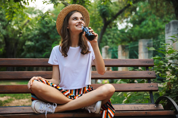 Happy young girl drinking from a bottle with fizzy drink while sitting at the park outdoors - Фото, зображення