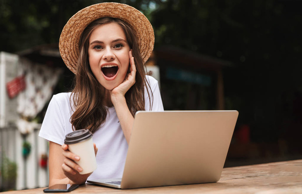 Cheerful young girl drinking coffee while sitting with laptop computer at the cafe outdoors - Foto, Bild