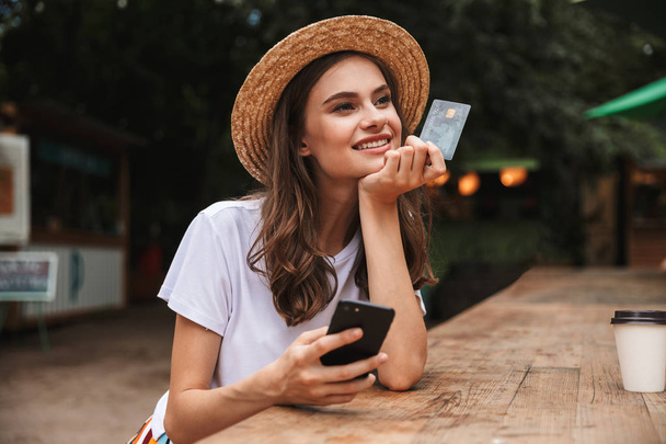 Happy young girl holding plastic credit card while using mobile phone at the cafe outdoors - Foto, imagen
