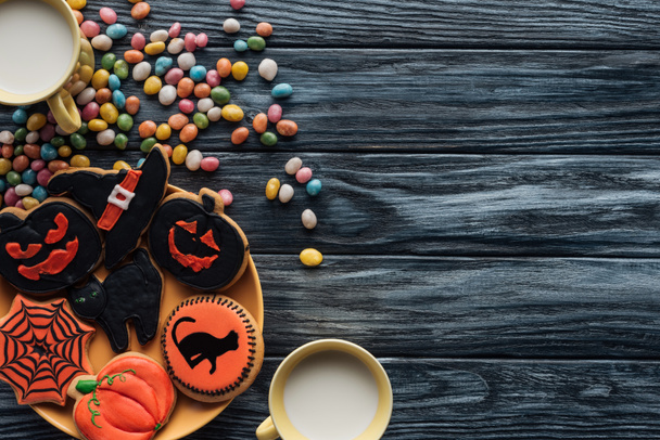 top view of plate with halloween cookies, colorful candies, cups with milk on wooden table  - Photo, Image