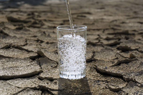 agua dulce pura y clara se vierte en un vaso de precipitados de vidrio de pie en medio de una tierra seca y desértica de arcilla agrietada
 - Foto, Imagen