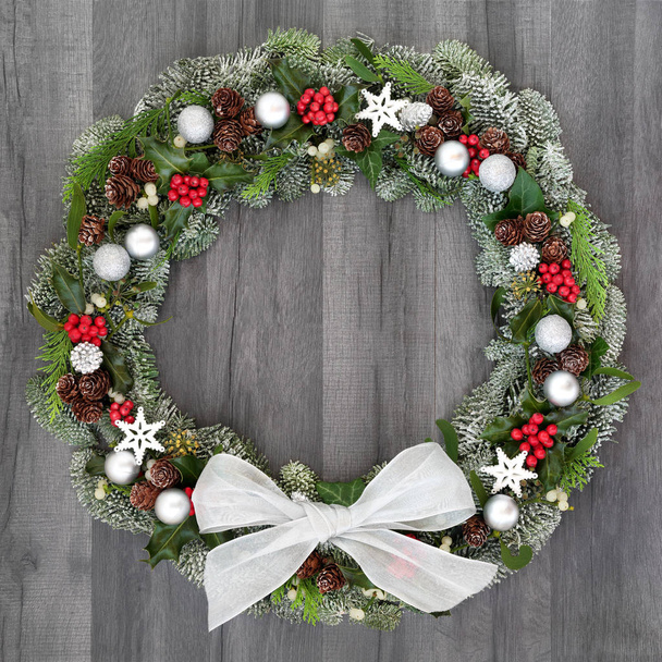 Corona de Navidad con flora invernal de acebo, muérdago, hiedra, abeto cubierto de nieve y conos de pino sobre fondo rústico de madera gris
. - Foto, imagen