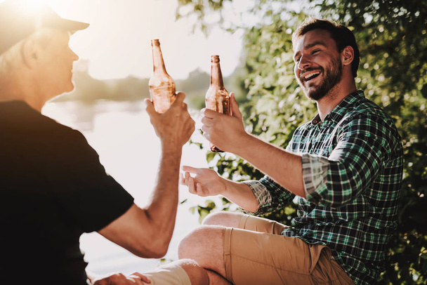 Smiling Men Drinking Beer near River in Summer. Man in Shirt. Bearded Guy. Weekend on River. Young Man. Relaxing Outdoor. Sitting Man. Man near Lake. Drinking Alkohol. Beer in Bottle. - Foto, Imagen