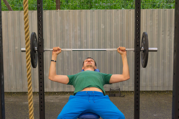 Un hombre con una camiseta verde y pantalones azules haciendo una barra de press de banco, sosteniendo la mancuerna en los codos, acostado en el gimnasio al aire libre en el fondo de una cuerda, una valla de metal y arbustos verdes
. - Foto, Imagen