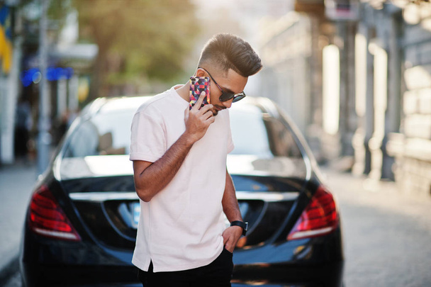 Stylish indian beard man at sunglasses and pink t-shirt against luxury car and speaking on mobile phone. India rich model posed outdoor at streets of city. - Photo, Image