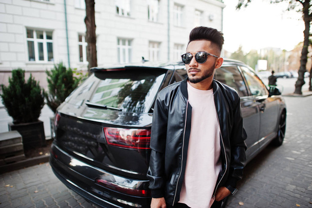 Stylish indian beard man at black leather jacket and sunglasses against business suv car. India model posed outdoor at streets of city. - Foto, Imagem