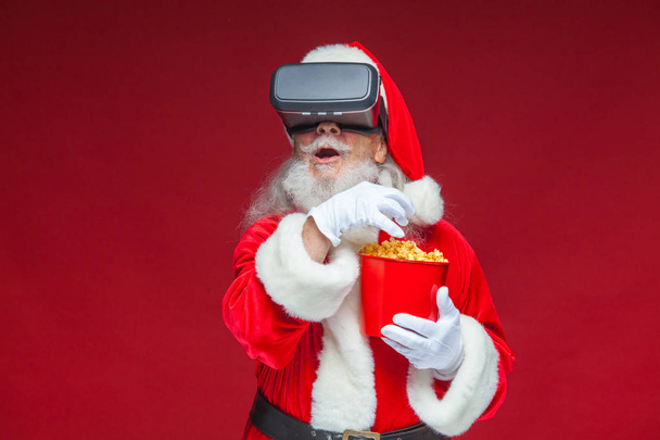 Christmas. Santa Claus in black virtual reality glasses holding a red bucket of popcorn. Watching a movie in virtual reality. Isolated on red background. - Valokuva, kuva
