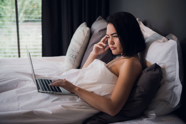 Young beautiful caucasian woman laying on bed and using laptop to work after wake up and thinking about work. Copy space - 写真・画像
