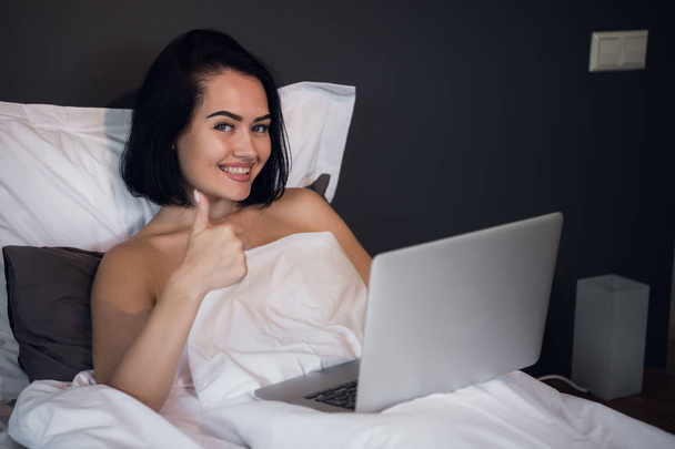 szczęśliwą kobietą, wpisując na swojego laptopa i pokazuje kciuk w górę leżąc w łóżku w domu - Zdjęcie, obraz