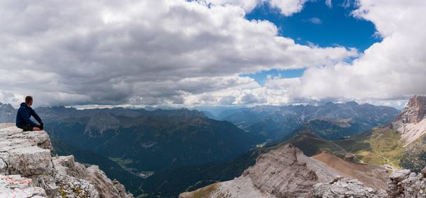 male hiker sitting on a mountain peak ledge in the Dolomites and looking at the amazing view of the surrounding landscape with Langkofel and Marmolada peaks - Photo, Image