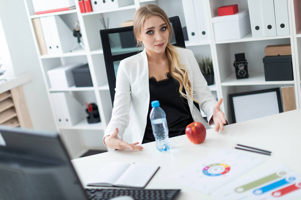 ein junges Mädchen sitzt an einem Tisch im Büro und zeigt auf eine Flasche Wasser und einen Apfel. - Foto, Bild