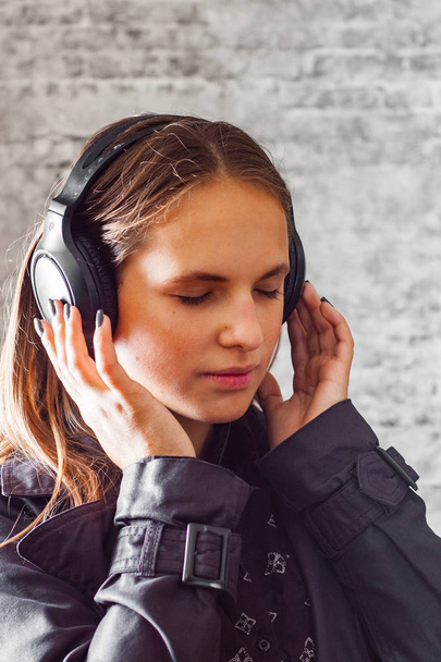 portrét mladé dospívající brunetka s dlouhými vlasy poslech hudby ve sluchátkách na pozadí šedé zdi - Fotografie, Obrázek