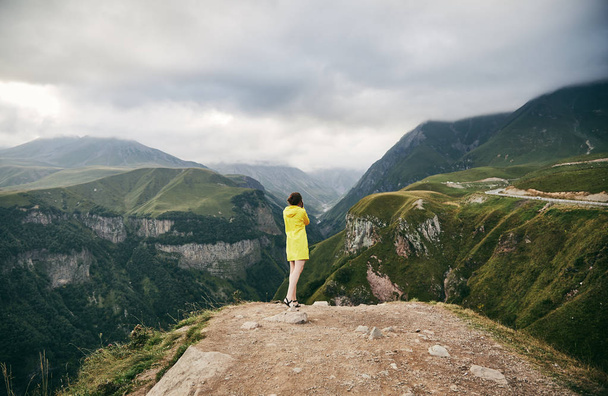 Ένα νεαρό κορίτσι σε ένα κίτρινο αδιάβροχο φωτογραφίζει τα βουνά. Γεωργία. Το καλοκαίρι. Αυγούστου. Κορίτσι κάνει μια φωτογράφηση του βουνού.  - Φωτογραφία, εικόνα